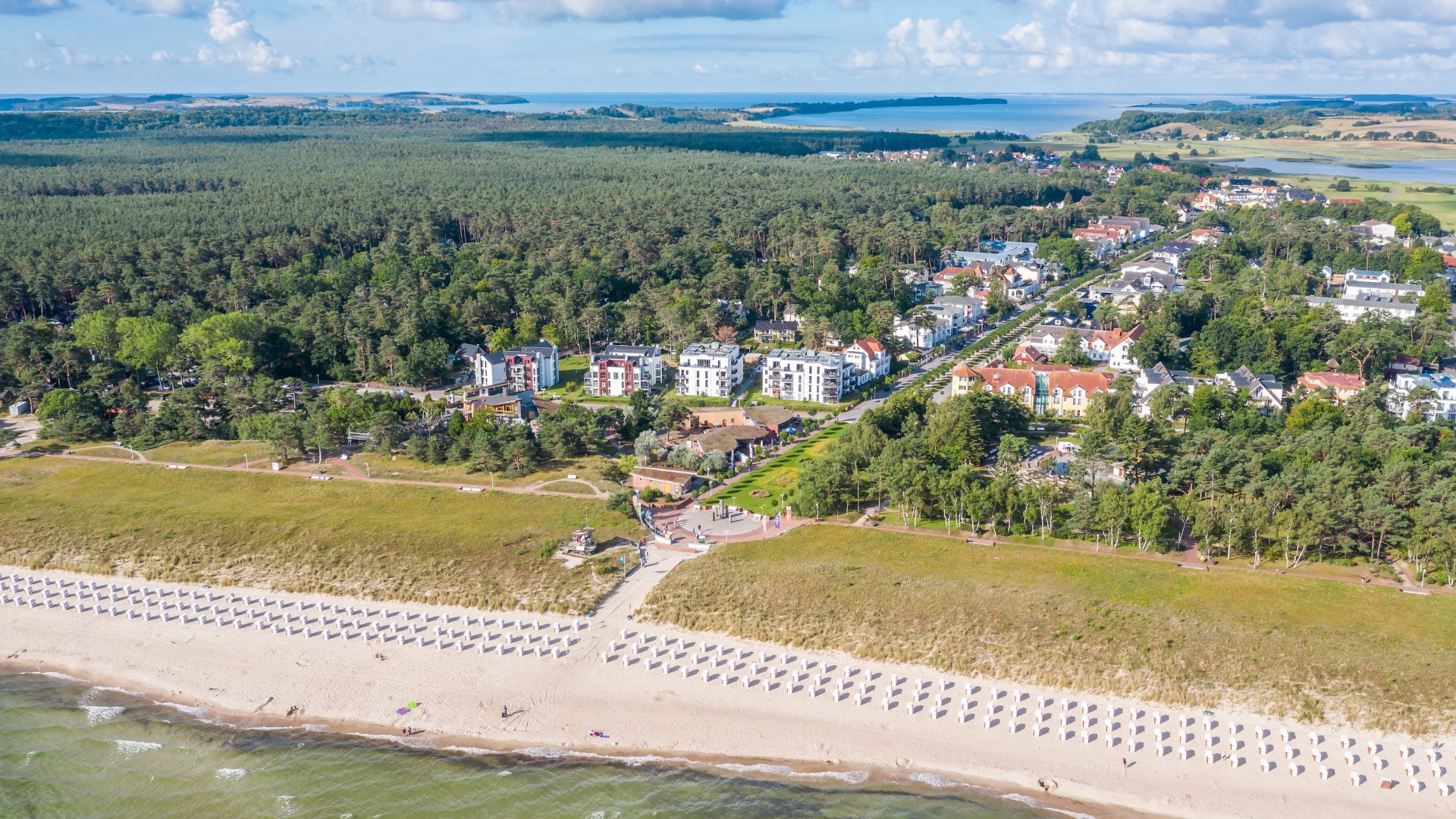 Aerial view of the Baltic seaside resort of Baabe on the island of R&uuml;gen: Beach with beach chairs, dunes, modern villas and vacation homes, surrounded by dense forests and views of the Bodden landscape.