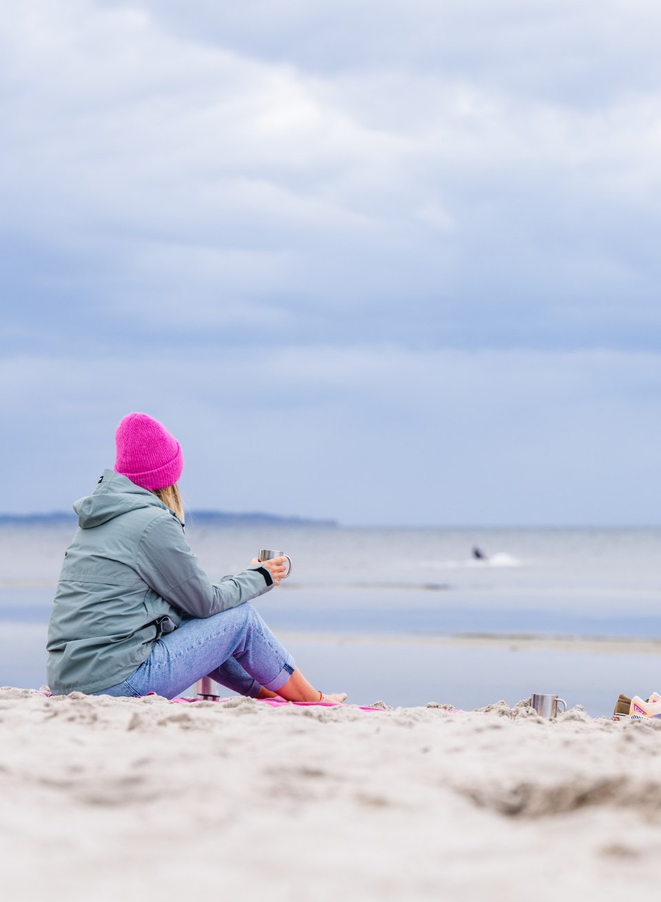  A woman in a pink cap is sitting on the beach in Timmendorf, looking out to sea and holding a cup in her hand.