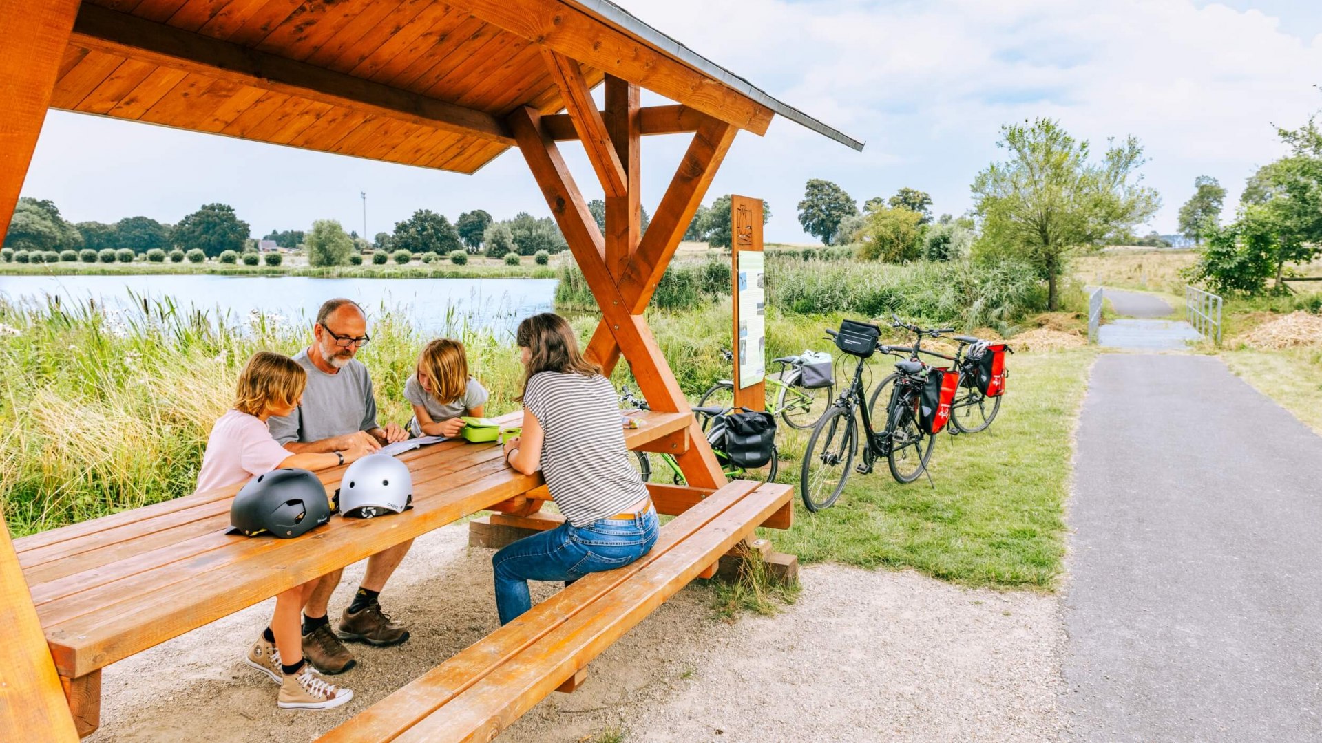 Een gezin neemt een pauze bij een rustplaats naast een meer na een fietstocht. // Zelfs de fitste fietsers hebben af en toe een pauze nodig. En het volgende meer in Mecklenburg glinstert altijd op de achtergrond. // © MV-T/Tiemann Een gezin neemt een pauze bij een rustplaats naast een meer na een fietstocht.