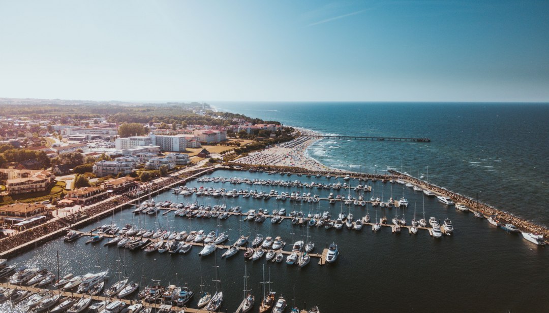 Widok z lotu ptaka na przystań Kühlungsborn z łodziami, plażą i Morzem Bałtyckim w promieniach słońca. // Przystań w Kühlungsborn leży między Morzem Bałtyckim a nadmorskim lasem. Łodzie kołyszą się w czystej wodzie, a plaża zachęca do kąpieli. Morski styl spotyka się z ogromem - to miejsce, do którego można przybyć i wziąć głęboki oddech. // © MV-T/Friedrich Widok z lotu ptaka na przystań Kühlungsborn z łodziami, plażą i Morzem Bałtyckim w promieniach słońca.