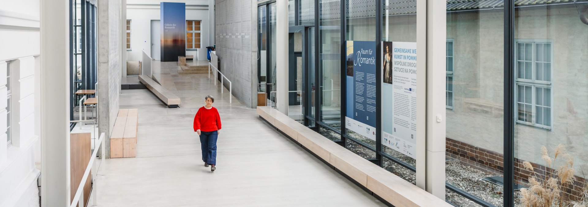 W Pomorskim Muzeum Państwowym w Greifswaldzie stare i nowe łączą się, tworząc imponujący zespół. Zalane światłem korytarze łączą historyczne części budynku ze współczesną architekturą i tworzą przestrzeń dla sztuki i spotkań. Miejsce, które łączy tradycję i nowoczesność., © MV-T/Gross Jasny korytarz muzealny ze szklaną fasadą w Pomorskim Muzeum Państwowym w Greifswaldzie, zwiedzający w czerwonym swetrze.