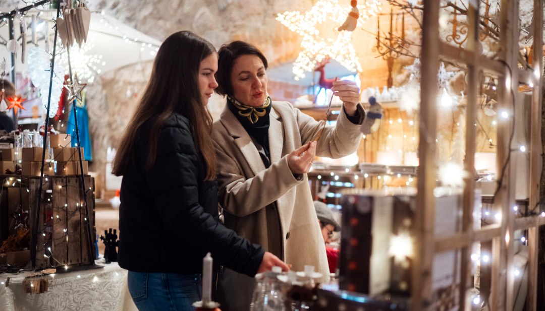 Two women at a festively decorated Christmas market look at lovingly designed decorations, surrounded by twinkling lights and a wintery atmosphere.