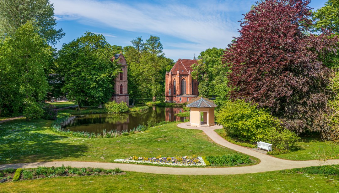 Het eerste neogotische kerkgebouw in Mecklenburg, een enkele klokkentoren en het oudste parkgebouw: verschillende hoogtepunten van Paleistuin Ludwigslust zijn te vinden in de Prinzengarten, direct naast het paleis., © TMV/Tiemann Klokkentoren en parkgebouw in de paleistuinen van Ludwigslust met bloemen en bomen.