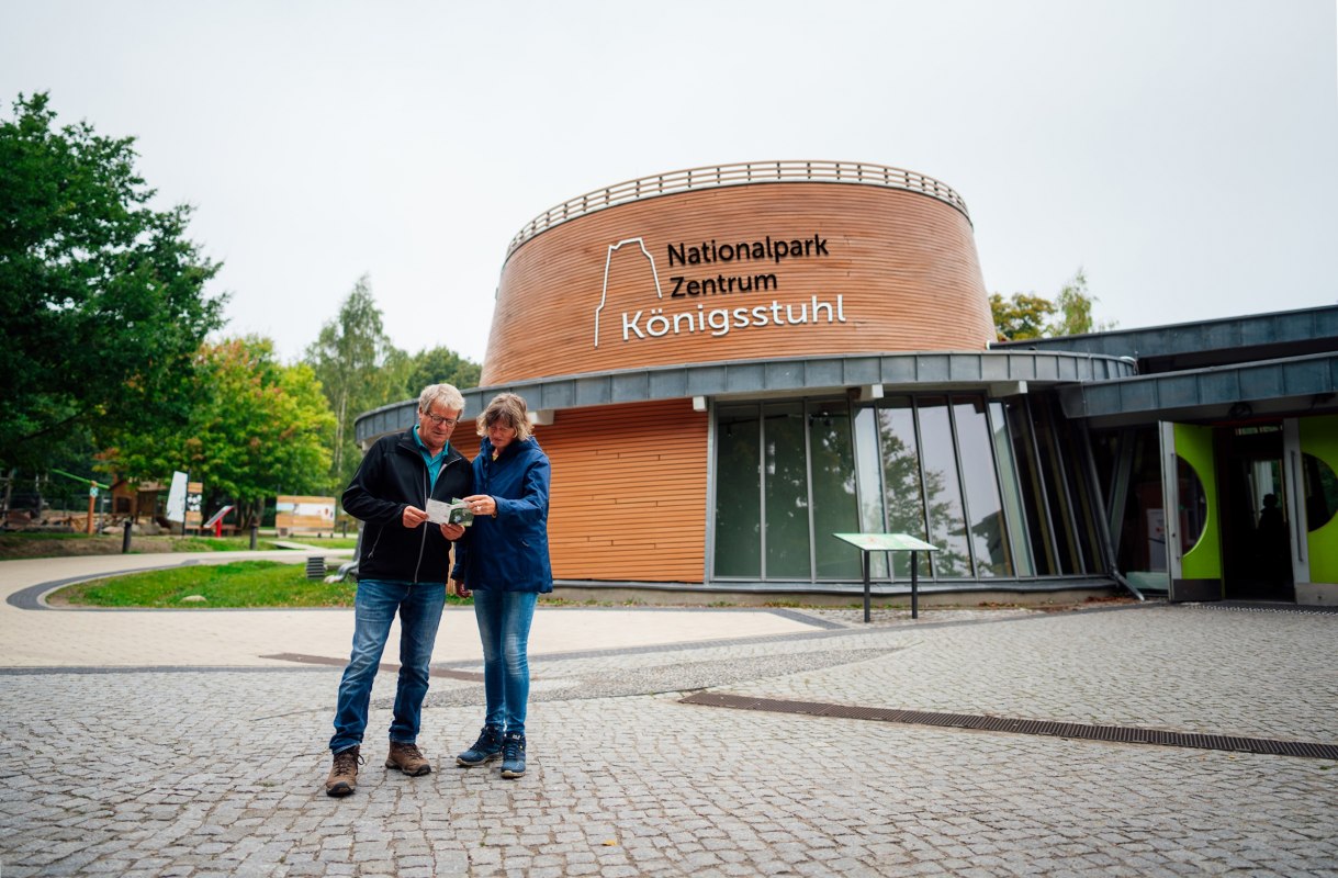 Nowoczesny budynek z okrągłym wejściem, przed kt&oacute;rym na wybrukowanym placu stoją dwie osoby. // &copy; Nationalpark-Zentrum K&Ouml;NIGSSTUHL / Dan Petermann