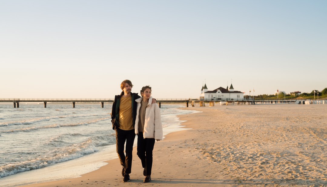 Para spacerująca po plaży Ahlbeck o wschodzie słońca z molo w tle, © TMV/Petermann Para spacerująca po plaży Ahlbeck o wschodzie słońca z molo w tle.