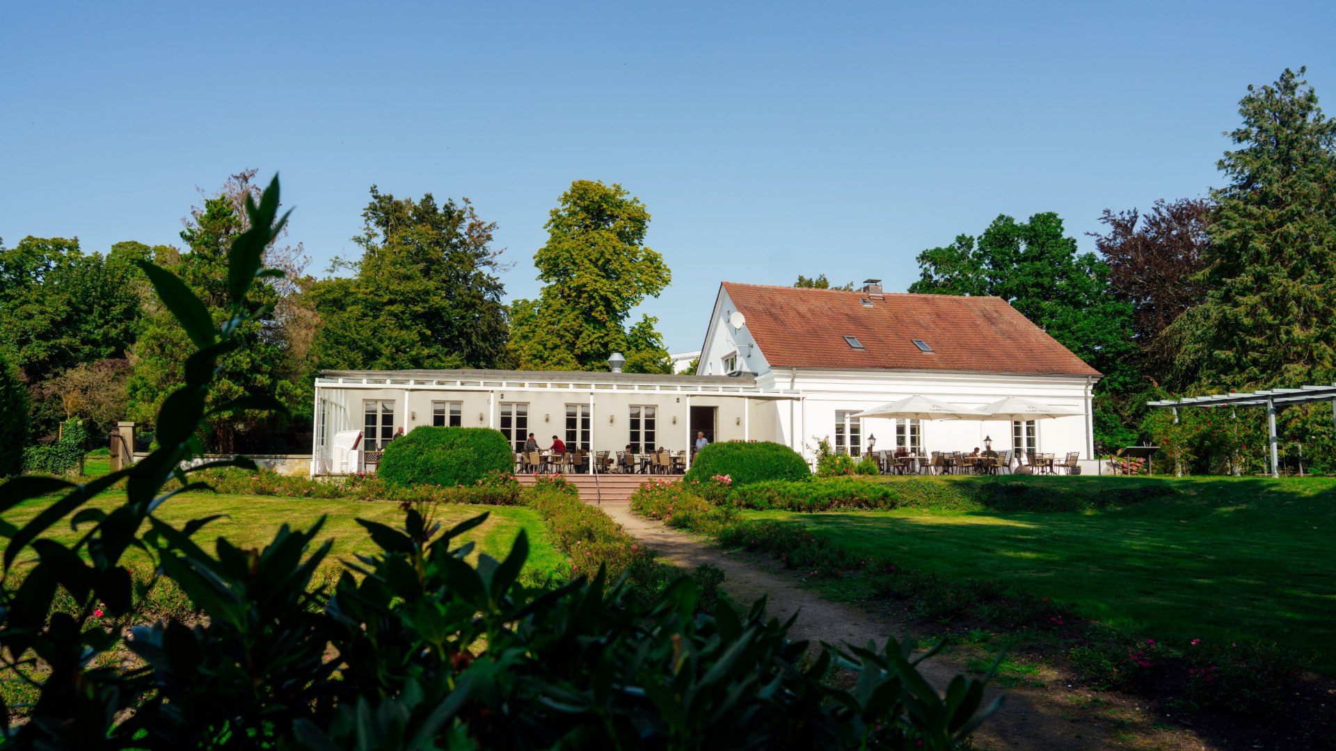 Het Rosencafé in Putbus op het eiland Rügen is een wit gebouw met een rood dak, omgeven door goed onderhouden tuinen en groene struiken. Tafels op het terras voor het café nodigen uit om te ontspannen in de buitenlucht. Op de achtergrond torenen hoge bomen de blauwe lucht in.
