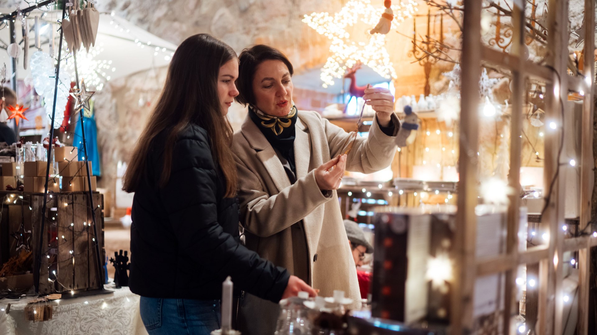 Two women at a festively decorated Christmas market look at lovingly designed decorations, surrounded by twinkling lights and a wintery atmosphere.