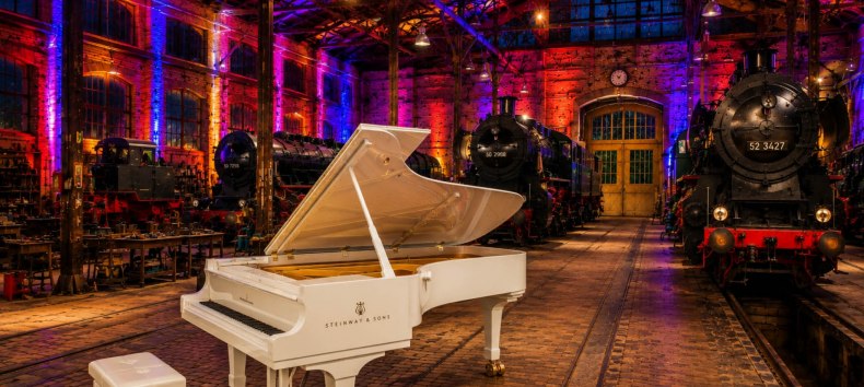 A large white grand piano in an old locomotive shed, with a steam locomotive in the background and atmospheric lighting // &copy; Tourismus, Freizeit & Kultur GmbH K&uuml;hlungsborn