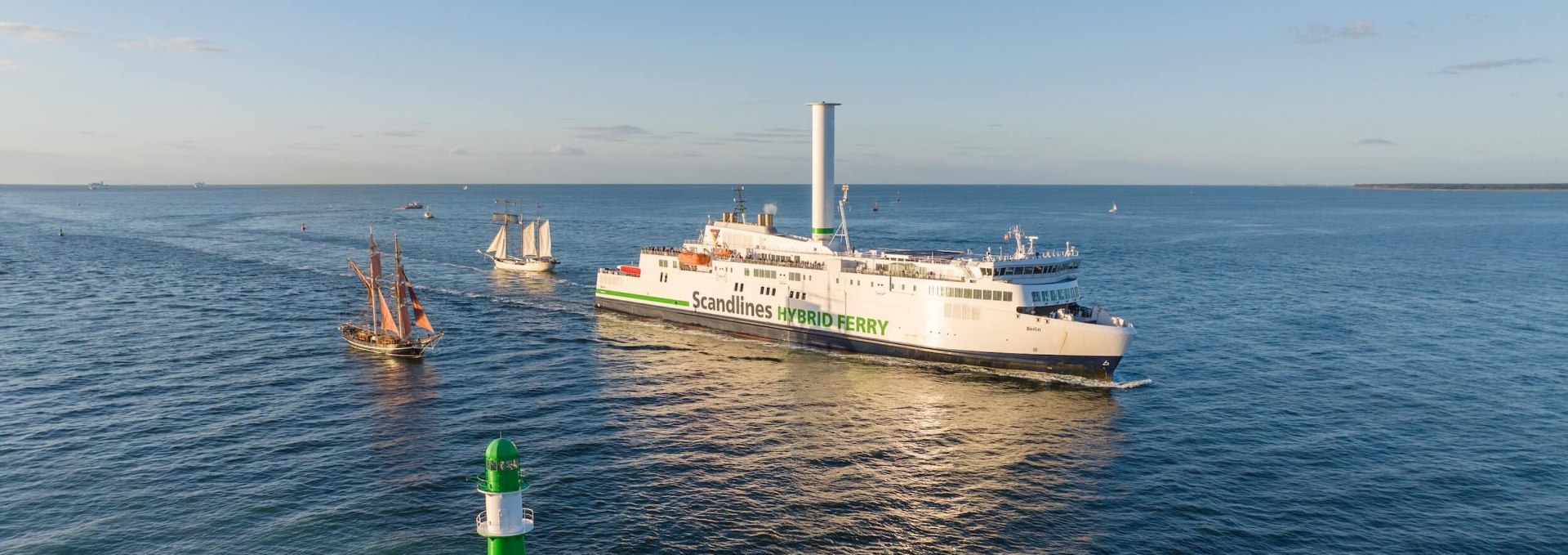 Hybrid ferry and tall ships sail past the green lighthouse at Warnemünde’s harbor entrance on the Baltic Sea.