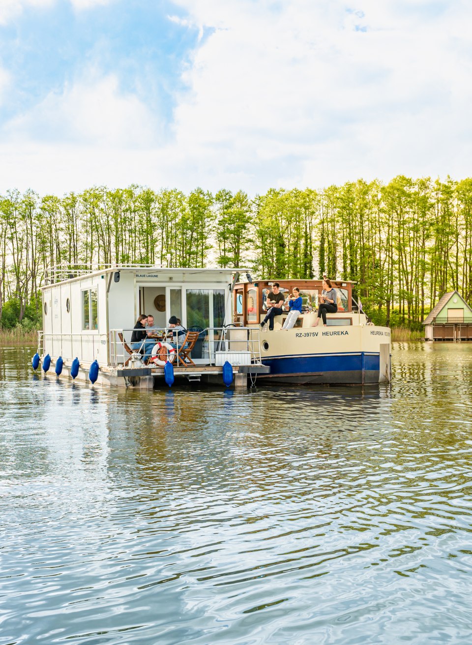 Although each of the two families has their own houseboat, they spend almost every minute of their vacation together. So do the cake breaks, for which they lay their boats next to each other., © TMV/Tiemann Two houseboats are moored next to each other, each with a family eating cake.