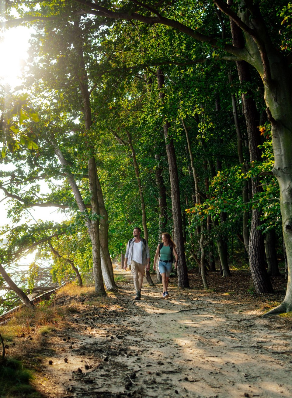 Een stel wandelt over een schaduwrijk bospad langs de kust in het biosfeerreservaat zuidoost R&uuml;gen. De zon schijnt door de bomen en wordt weerspiegeld in het water. De sfeer is rustig en wordt gekenmerkt door de natuurlijke schoonheid van de omgeving.