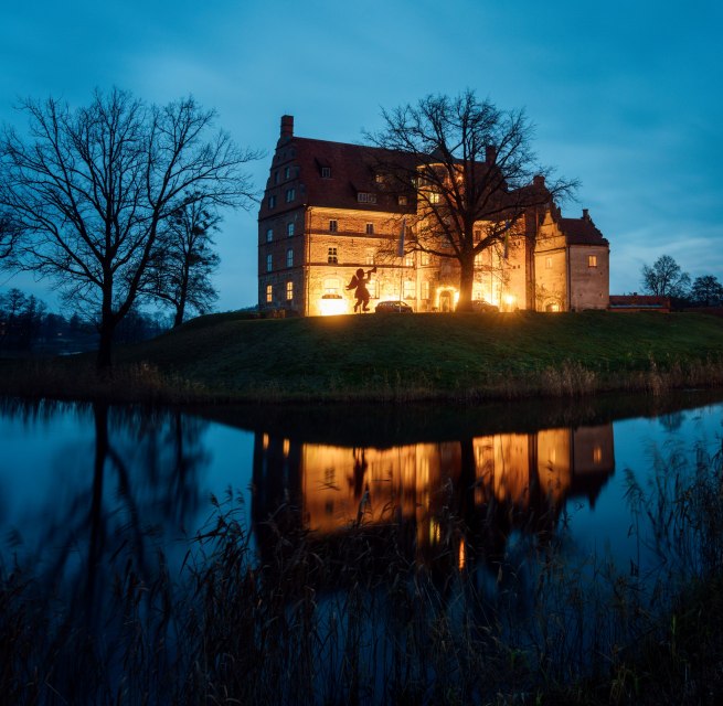 Atmospherically illuminated Ulrichshusen Castle at Christmas time, surrounded by trees and a moat with reflections in the evening light.