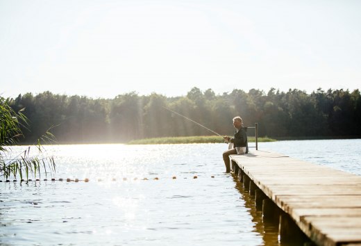 Een oudere man zit op een houten steiger aan een rustig meer en vist terwijl de zon het water laat glinsteren.