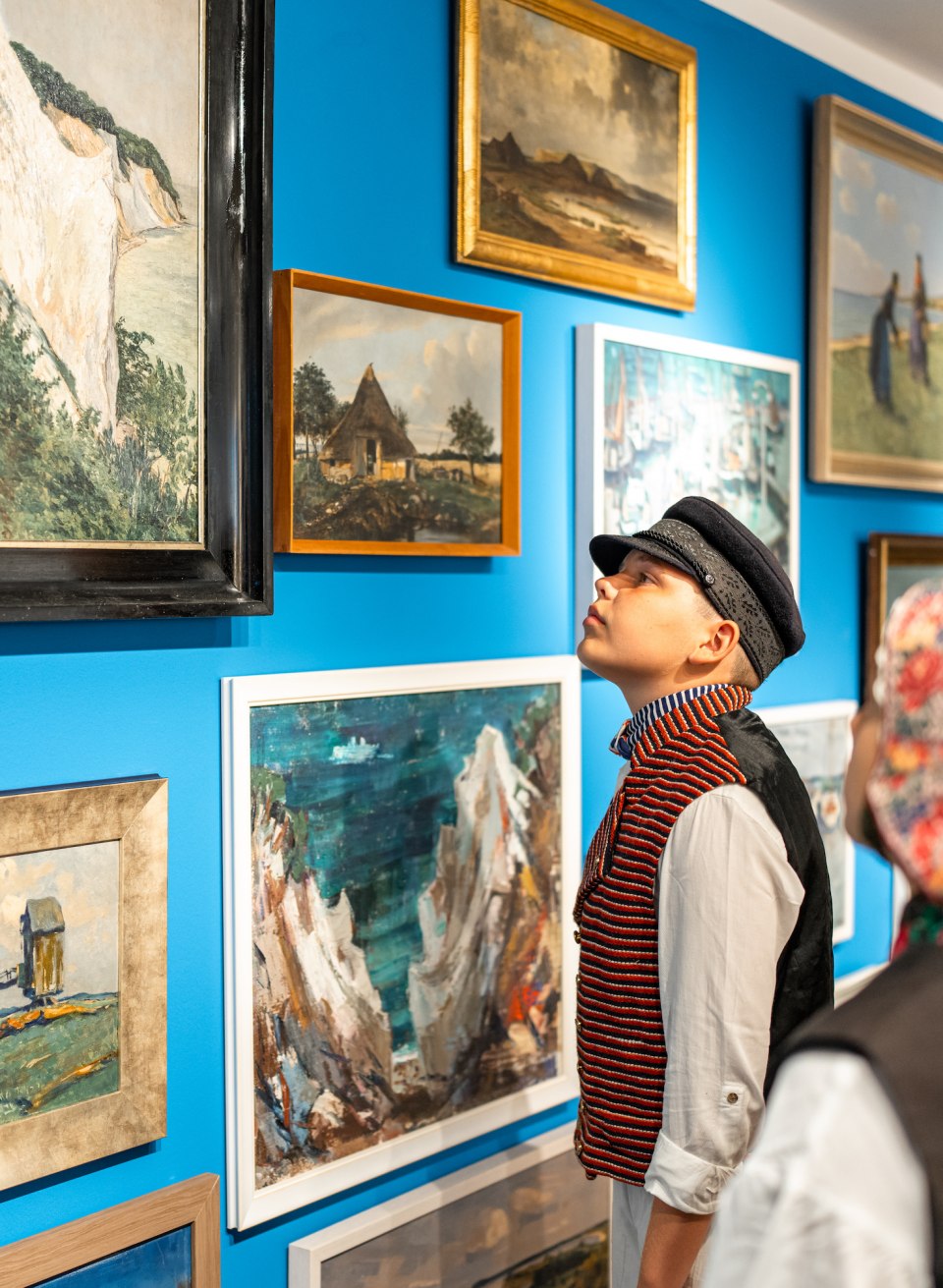 Drawings and paintings hang on the wall in the M&ouml;nchgut local history museum - children stand in front of it in traditional dress and examine them.