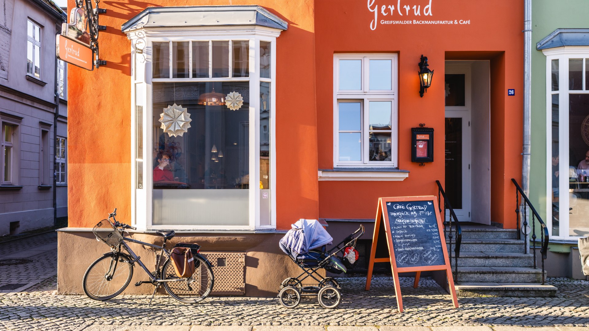 Oranjekleurige gevel van Caf&eacute; Gertrud met een witte erker in de oude binnenstad van Greifswald, met een fiets en kinderwagen ervoor.