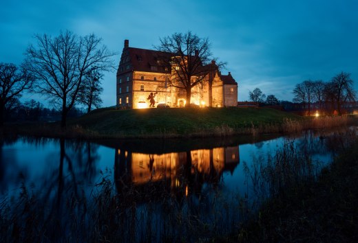 Atmospherically illuminated Ulrichshusen Castle at Christmas time, surrounded by trees and a moat with reflections in the evening light.