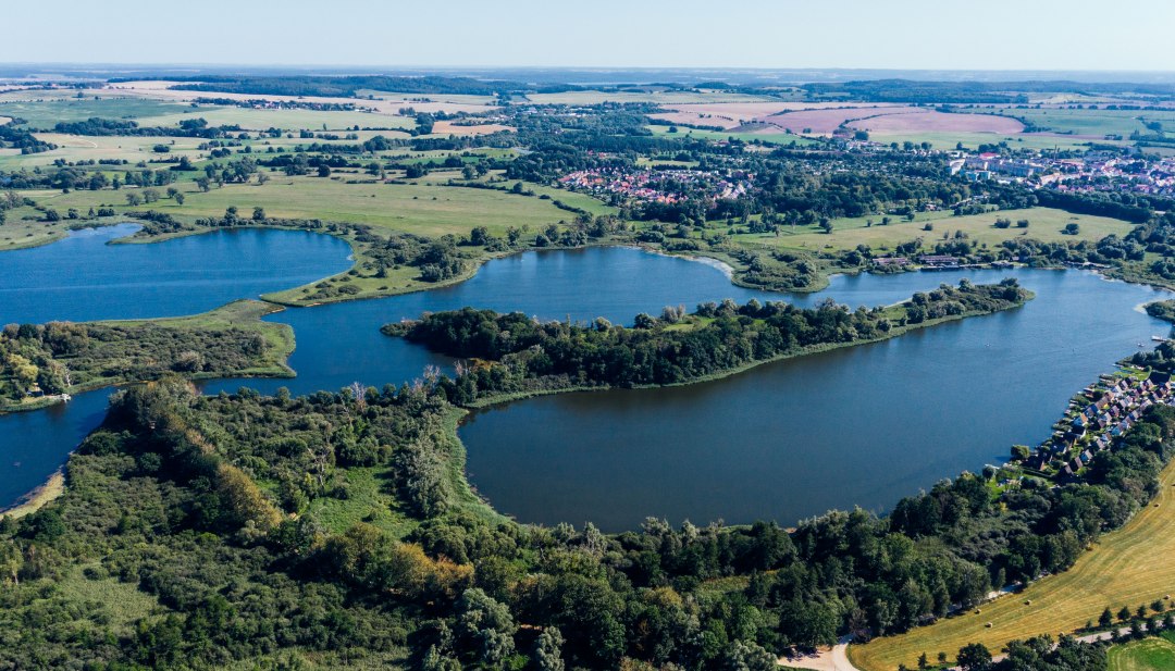 Widok z lotu ptaka na jezioro Teterow z Burgwallinsel, zielonymi brzegami, polami i Teterow w tle. // Rozległe przestrzenie wodne i zielone brzegi otaczają Burgwallinsel w jeziorze Teterow - przestrzeń do wzięcia głębokiego oddechu i odkrywania w Meklemburgii-Pomorzu Przednim. // © MV-T/Gänsicke Widok z lotu ptaka na jezioro Teterow z Burgwallinsel, zielonymi brzegami, polami i Teterow w tle.