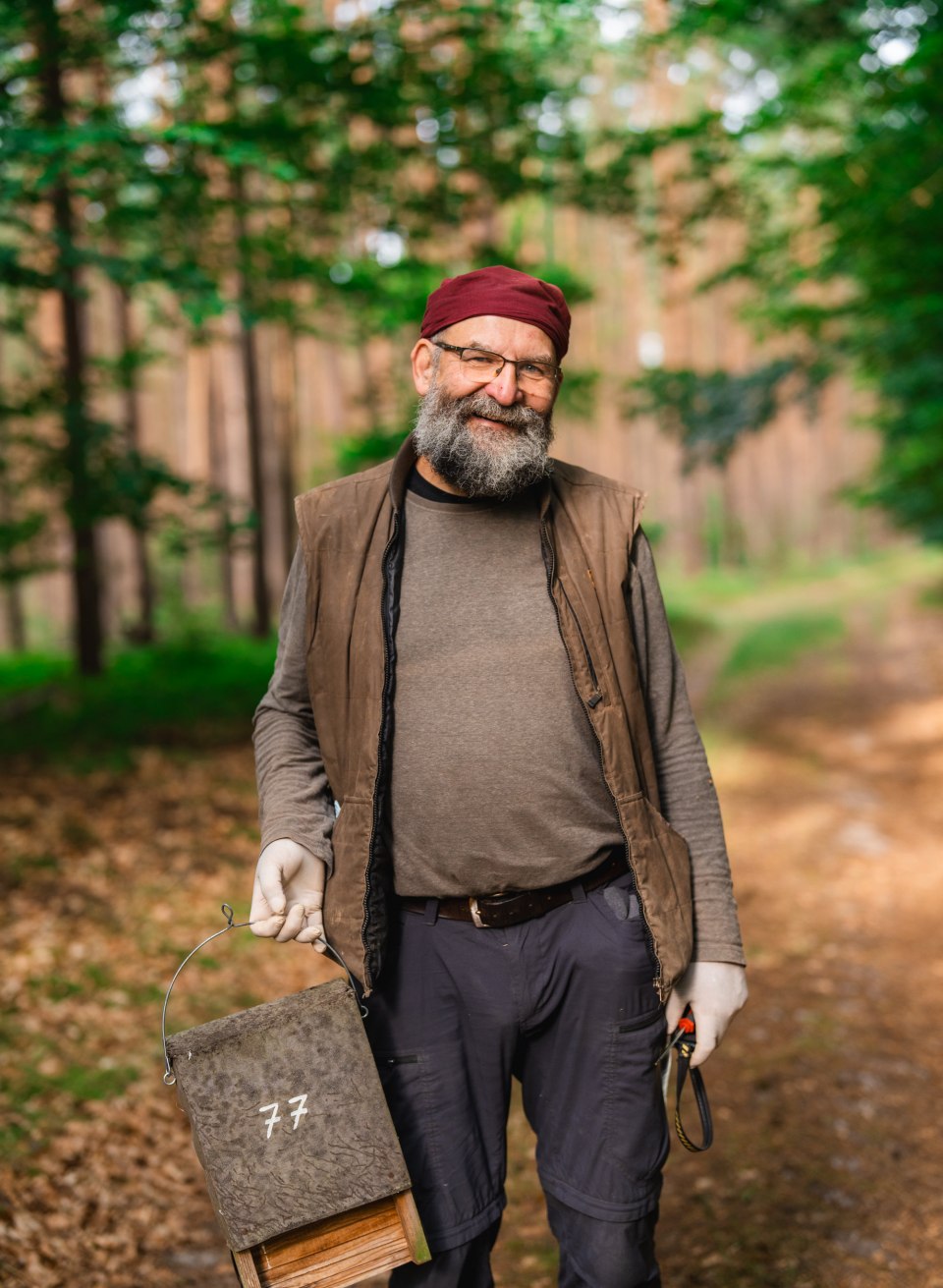 A man is standing on a forest path, holding a numbered bat box in his hand, with trees in the background.