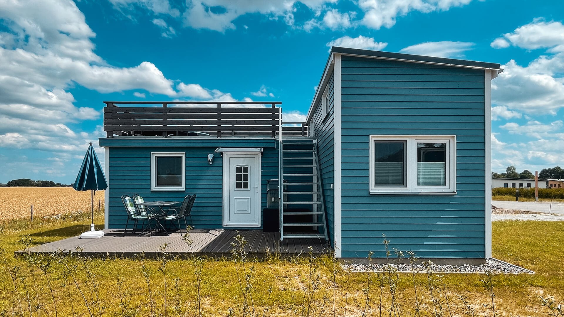 Ontspan in een idyllische omgeving - stijlvol Tiny House met dakterras en patio voor ontspannende uren in de natuur aan de Baltische Zee., © Prima Camping Resort Boddenblick Modern, blauw Tiny House met dakterras en gezellig houten terras met zitje, omgeven door landelijk landschap onder een blauwe hemel.
