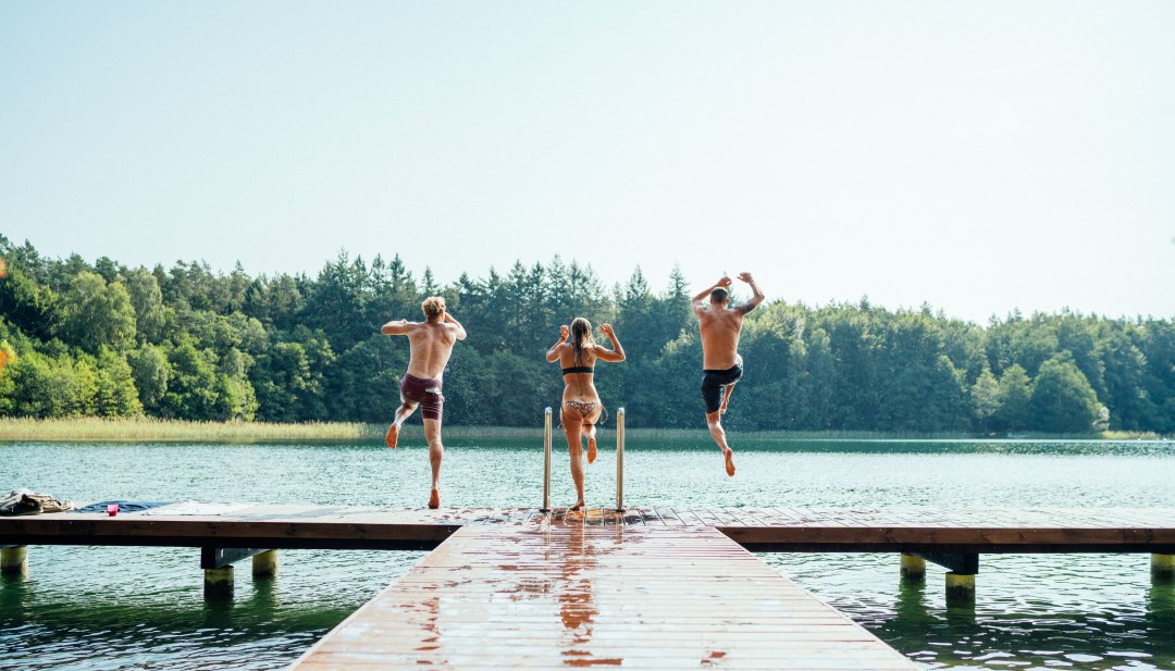 Drie jonge mensen springen samen van een houten loopbrug in een helder meer omringd door dichte bossen.
