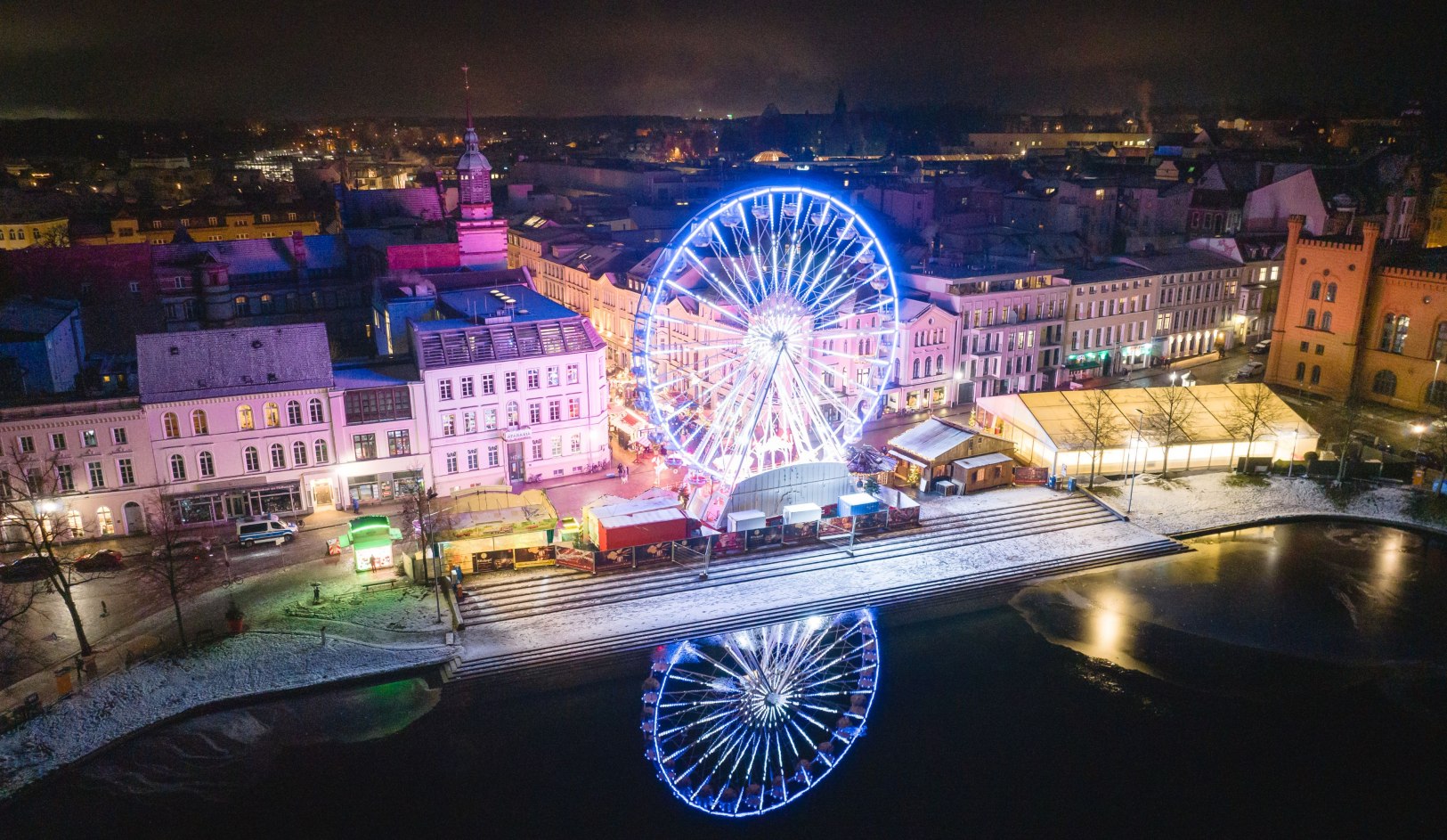 e glamour van de Pfaffenteich - het reuzenrad op de kerstmarkt van Schwerin biedt onvergetelijke uitzichten in een feestelijke sfeer., © Erik Gross Verlicht reuzenrad bij de Pfaffenteich vijver in Schwerin met winterse kerstmarkt en weerspiegeling in het water bij nacht.