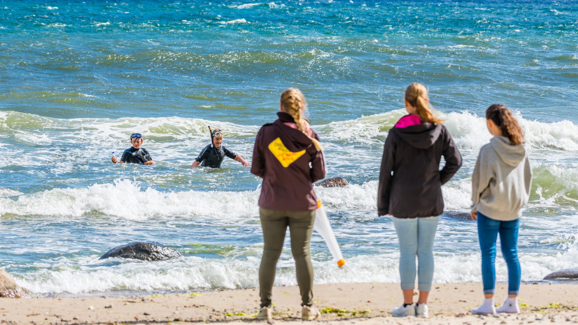 Bemonstering in de Oostzee Göhren - twee studenten in wetsuit komen uit de Oostzee naar de wachtende mensen op het strand. // Twee van de jonge rangers in wetsuits en met planktonnetten nemen monsters uit de golvende Baltische Zee. // © MV-T/Tiemann Bemonstering in de Oostzee Göhren - twee studenten in wetsuit komen uit de Oostzee naar de wachtende mensen op het strand.