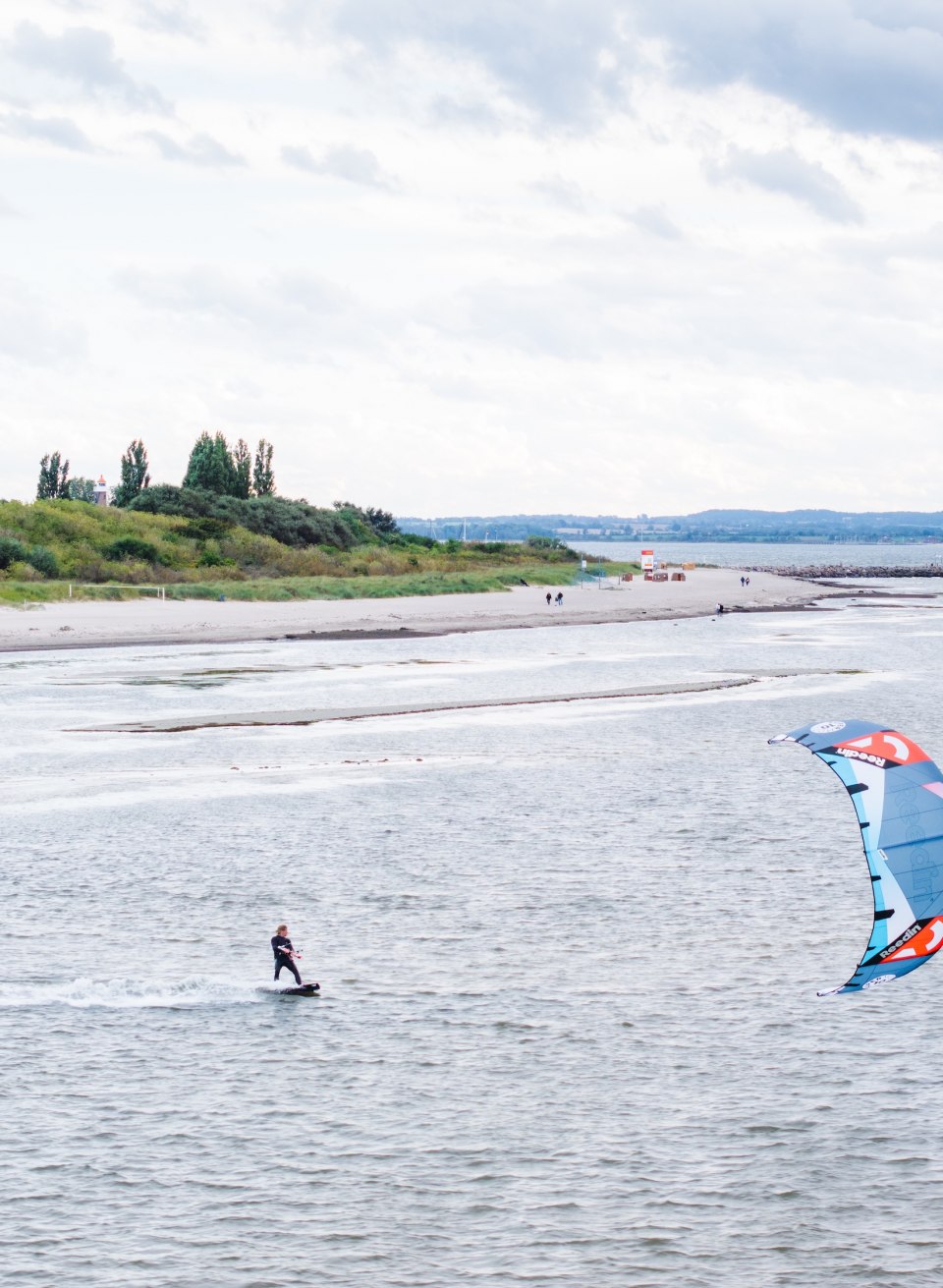 A kitesurfer glides over the water off the coast of Timmendorf on Poel, with a green dune landscape in the background.