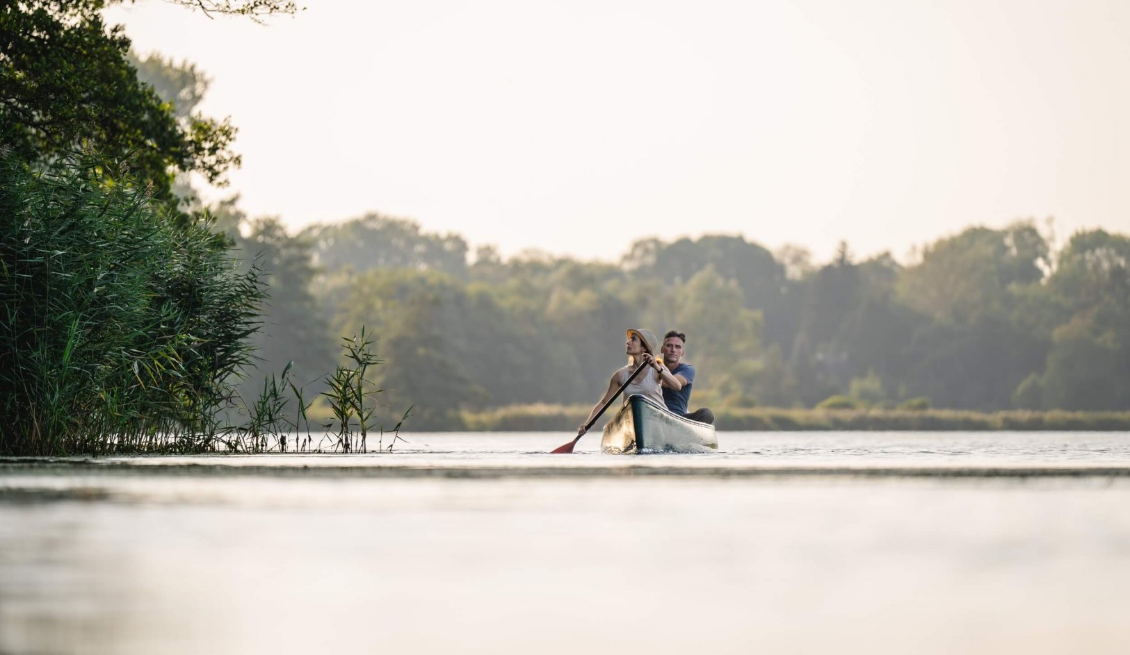 Een koppel peddelt samen in een kano over de rivier de Peene in Mecklenburg-Vorpommern, omringd door riet en bomen op de achtergrond.
