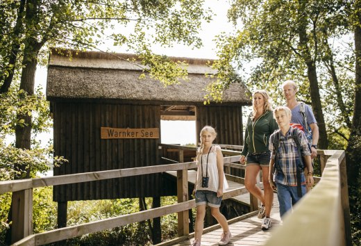 Eine Familie geht über einen Holzsteg zu einer Hütte mit Reetdach, ein Mädchen trägt ein Fernglas um den Hals. // Knarrende Holzbohlen, raschelndes Laub: Am Warnker See im Müritz-Nationalpark erkunden Familien den Steg zur Reetdachhütte. // © MV-T/Roth Eine Familie geht über einen Holzsteg zu einer Hütte mit Reetdach, ein Mädchen trägt ein Fernglas um den Hals. // Knarrende Holzbohlen, raschelndes Laub: Am Warnker See im Müritz-Nationalpark erkunden Familien den Steg zur Reetdachhütte. // © MV-T/Roth