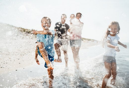A family runs through the shallow Baltic Sea waves on the beach at Dierhagen, laughing as water splashes vividly in the sun. // Warm sunlight, splashing waves and exuberant footsteps create a feeling of arrival on Dierhagen beach, inviting you to immerse yourself in the vast Baltic Sea summer. // © MV-T/Roth A family runs through the shallow Baltic Sea waves on the beach at Dierhagen, laughing as water splashes vividly in the sun.