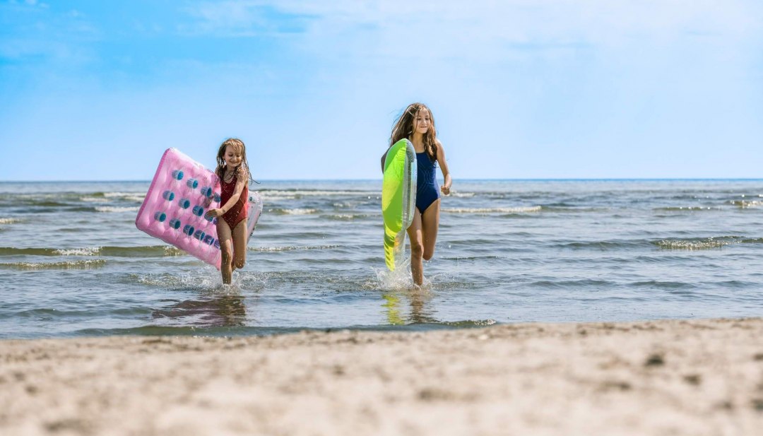 Dwoje dzieci wybiega z wody na plażę z dmuchanymi materacami pod pachami