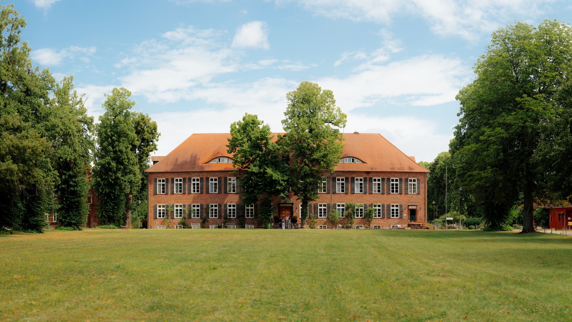 The Ludorf manor house, a brick building, stands behind a large meadow, surrounded by old trees under a blue sky. // A large meadow stretches out in front of the Ludorf manor house, with brick buildings and towering trees framing the setting—here, Jannis Achtenhagen focuses on tranquility, space, and the feeling of coming home in Mecklenburg-Western Pomerania. // © MV-T/Petermann The Ludorf manor house, a brick building, stands behind a large meadow, surrounded by old trees under a blue sky.
