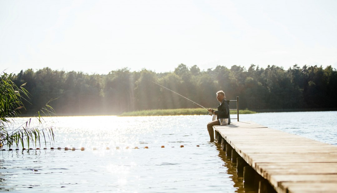 A man sits on a wooden jetty on a calm lake and fishes while the sun makes the water glisten.