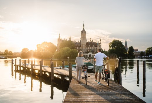 A couple with bicycles stand on a jetty on Lake Schwerin and look at Schwerin Castle in the evening light.