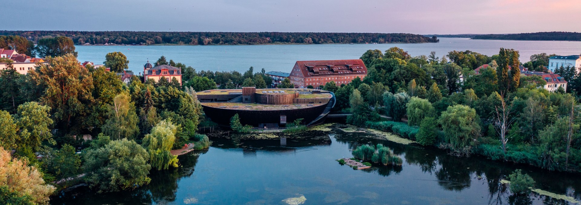 Luftfoto af M&uuml;ritzeum i Waren (M&uuml;ritz), omgivet af vand og gr&oslash;n natur i skumringen.