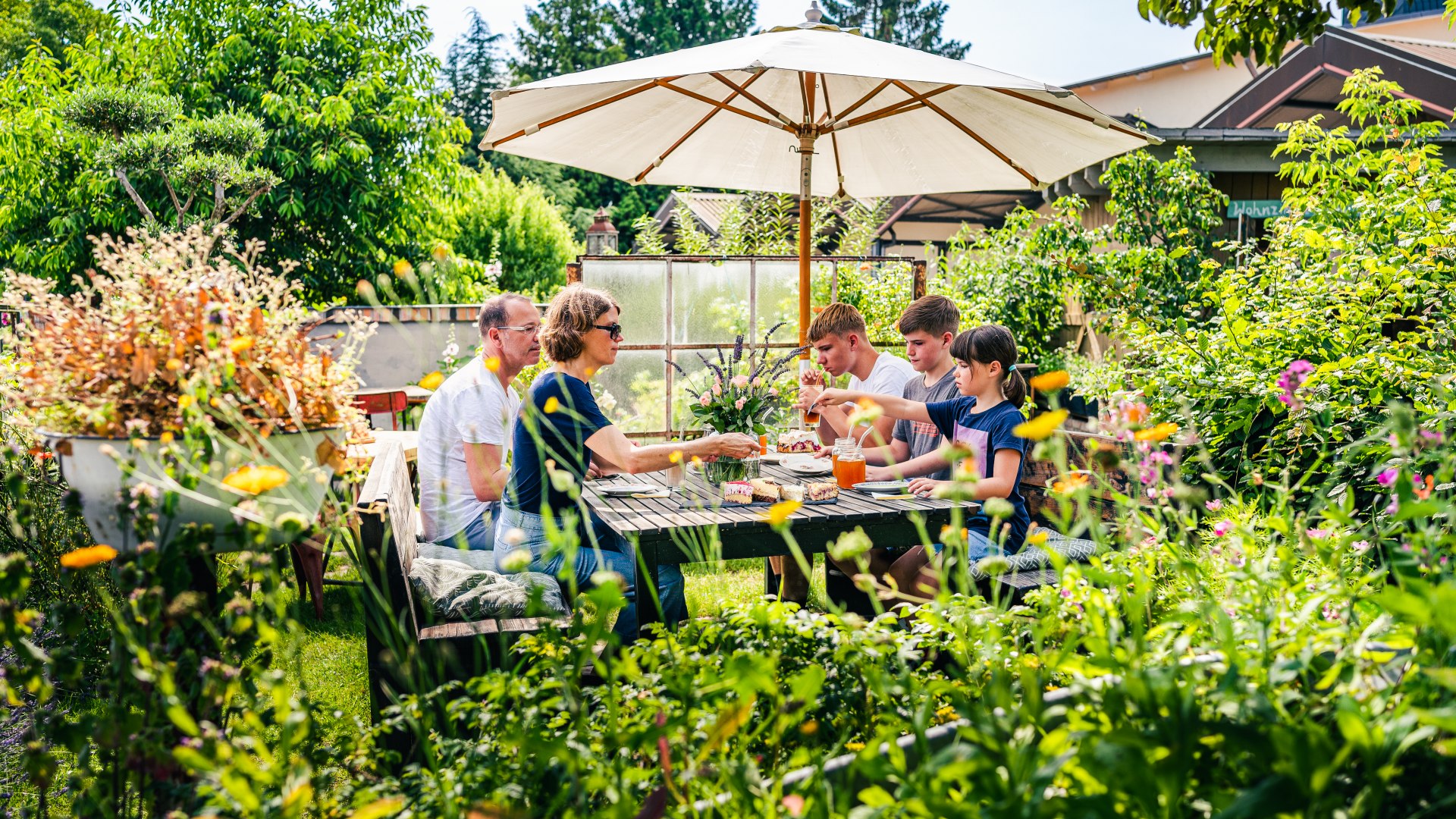Een gezin neemt een koffiepauze in de tuin van het café, die overwoekerd is met planten.