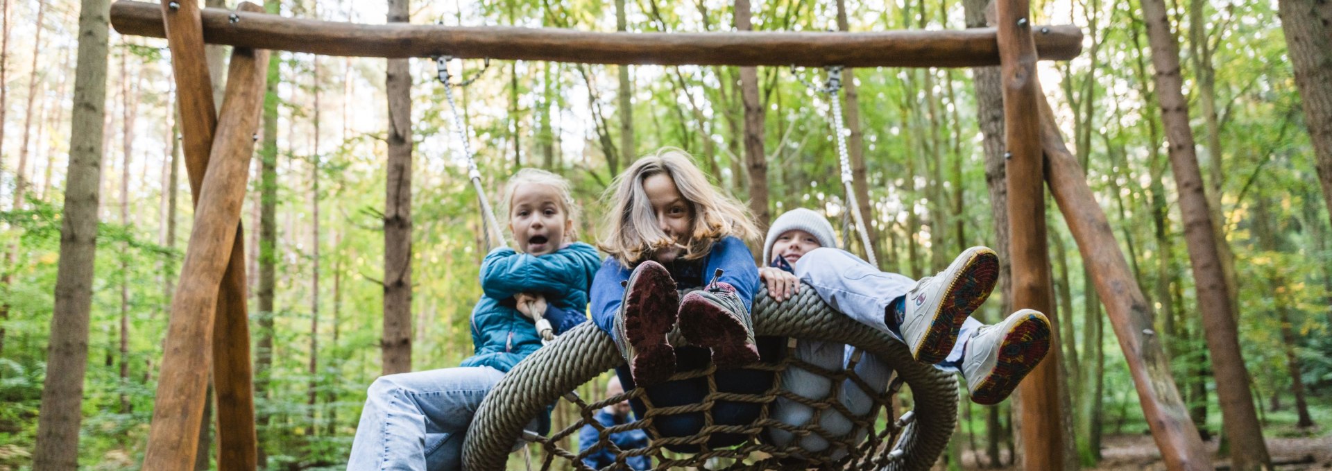 Drie kinderen zitten samen op een nestschommel van touwen in het bos en schommelen wild heen en weer.