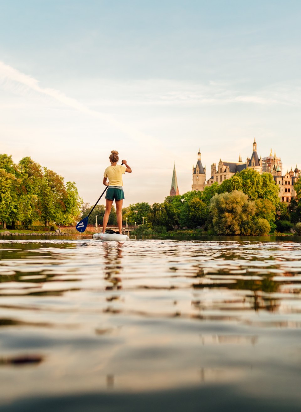 A person stands stand-up paddling on Lake Schwerin and paddles towards Schwerin Castle in the warm evening light.
