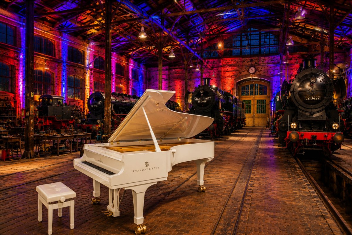 A large white grand piano in an old locomotive shed, with a steam locomotive in the background and atmospheric lighting // © Tourismus, Freizeit & Kultur GmbH Kühlungsborn A large white grand piano in an old locomotive shed, with a steam locomotive in the background and atmospheric lighting // © Tourismus, Freizeit & Kultur GmbH Kühlungsborn