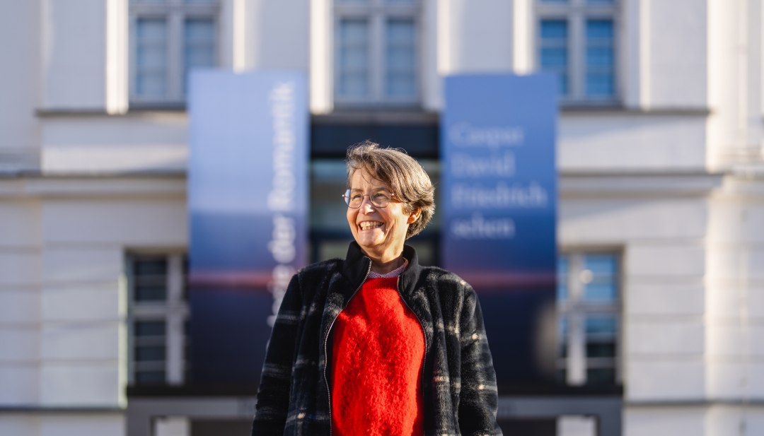 Kobieta w czerwonym swetrze stoi uśmiechnięta przed wejściem do Pomorskiego Muzeum Państwowego w Greifswaldzie.