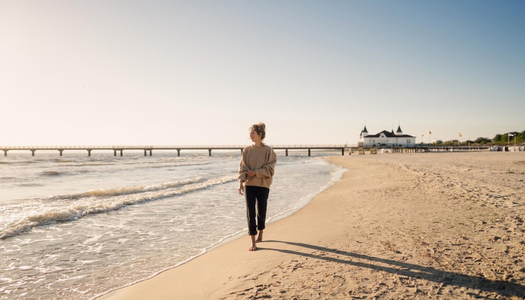 Kobieta spacerująca po plaży Ahlbeck o wschodzie słońca z molo w tle // © MV-T/Petermann Kobieta spacerująca po plaży Ahlbeck o wschodzie słońca z molo w tle