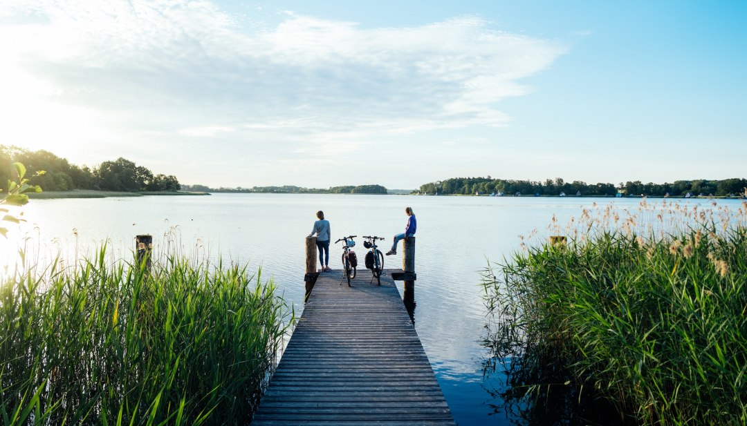 Two cyclists on a wooden footbridge on Krakow Lake surrounded by reeds and lake scenery.