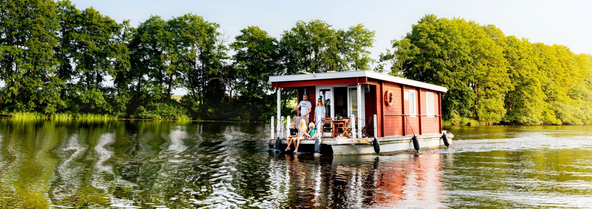Familie op een rode woonboot op een rustig meer, omringd door groene bomen en blauwe lucht.