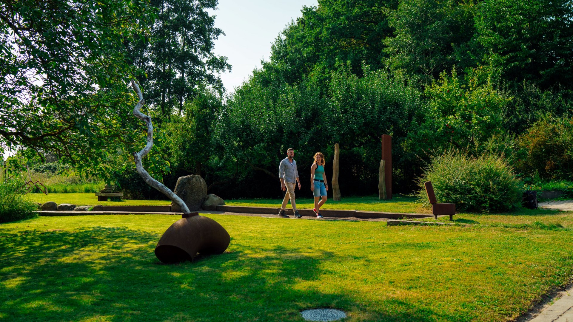Een koppel wandelt door een groene beeldentuin in de oude watermolen in Wreechen. Een moderne metalen kunstinstallatie staat op de voorgrond, terwijl bomen en struiken de tuin omlijsten. De rustige sfeer nodigt uit om te verkennen en te blijven hangen.