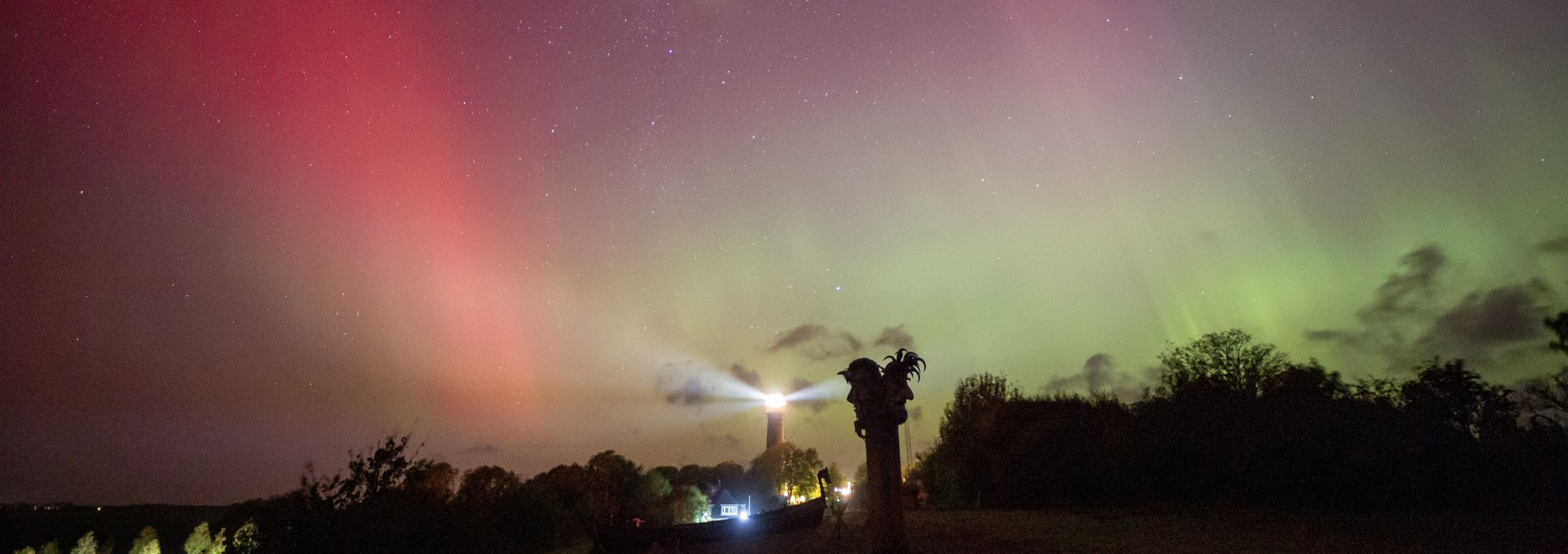 Noorderlicht in heldere rode en groene kleuren aan de nachtelijke hemel boven Kaap Arkona op het eiland Rügen, met de vuurtoren en een Vikingbeeldhouwwerk op de voorgrond.