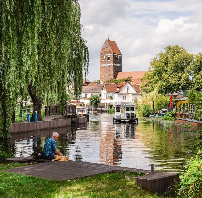 A man sits with his dog on the banks of a river in the center of Parchim, surrounded by willow trees and traditional houses in the background. // © MV-T/Gross A man sits with his dog on the banks of a river in the center of Parchim, surrounded by willow trees and traditional houses in the background.