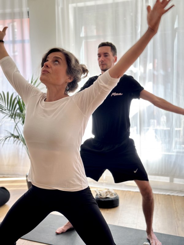 Twee mensen oefenen yoga in een helder verlichte studio. Op de voorgrond staat een vrouw in een krachtige staande yogahouding met haar armen omhoog, op de achtergrond volgt een man dezelfde beweging. Zacht daglicht en een kalme sfeer benadrukken mindfulness, beweging en lichaamsbewustzijn., © MOVA PLACE - Yoga & Co. Studio Twee mensen oefenen yoga in een helder verlichte studio. Op de voorgrond staat een vrouw in een krachtige staande yogahouding met haar armen omhoog, op de achtergrond volgt een man dezelfde beweging. Zacht daglicht en een kalme sfeer benadrukken mindfulness, beweging en lichaamsbewustzijn., © MOVA PLACE - Yoga & Co. Studio