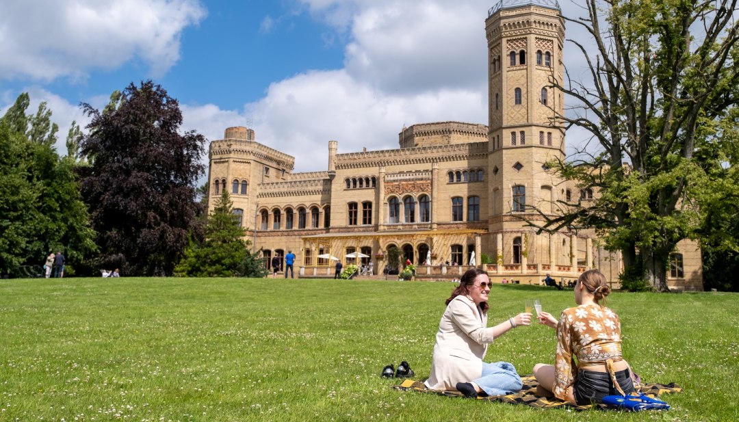 Two women enjoy a picnic on a meadow in front of Neetzow Castle on a sunny day during the Midsummer Remise.