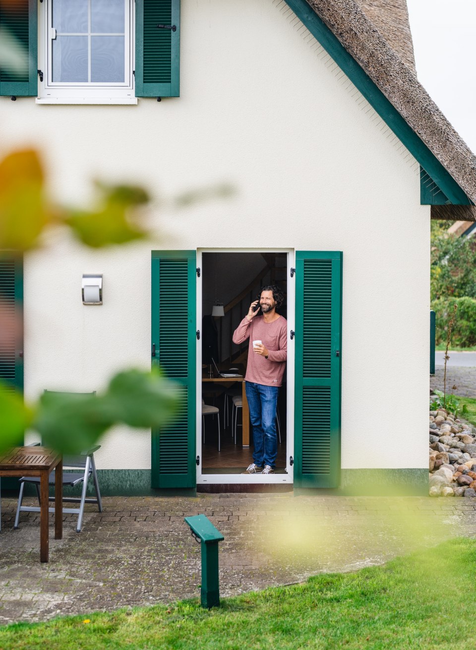 A man talks on the phone and drinks coffee in the doorway of a vacation home with green shutters on the island of Poel.