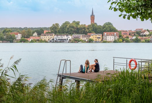 Two women sit on a jetty and look out over a lake